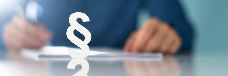 Ein Paragraphensymbol im Vordergrund, sieht man unscharf im Hintergrund einen Mann ein Dokument überarbeitend